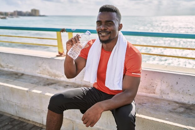 Jonge afro-amerikaanse man met sportkleding die waterfles vasthoudt aan zee