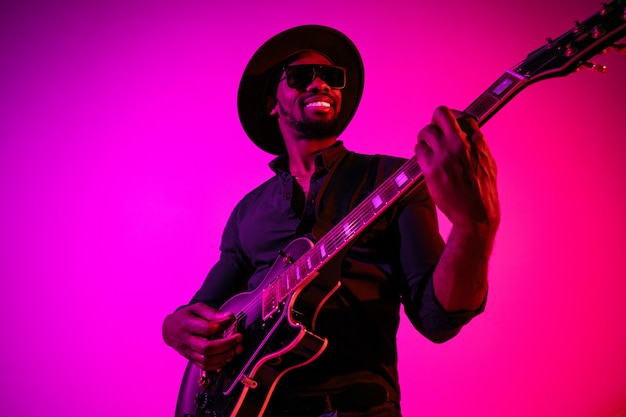 Gratis foto jonge afro-amerikaanse muzikant die gitaar speelt als een rockster op een paarsroze gradiëntmuur in neonlicht