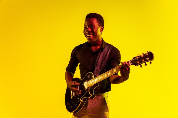 Jonge afro-amerikaanse muzikant die gitaar speelt als een rockster op gele achtergrond in neonlicht. Concept van muziek, hobby, festival, openlucht. Vrolijke man improviseren, zingen lied.