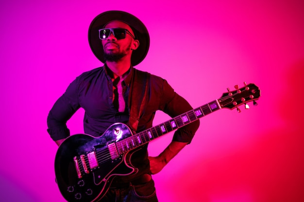 Jonge Afro-Amerikaanse muzikant gitaar spelen als een rockstar op paars-roze achtergrond met kleurovergang in neonlicht.