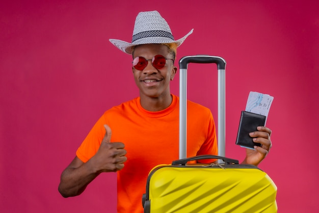 Jonge Afro-Amerikaanse reiziger jongen dragen oranje t-shirt en zomer hoed houden reiskoffer en vliegtickets kijken camera glimlachend positief en gelukkig duimen opdagen op roze achtergrond