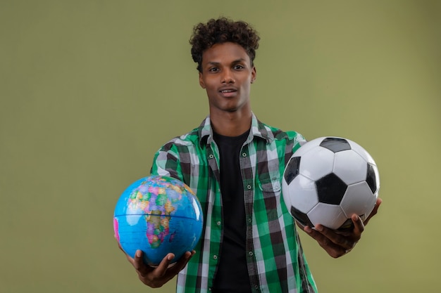 Jonge afro-amerikaanse reiziger man met globe en voetbal kijken camera glimlachend blij en positief staande over groene achtergrond