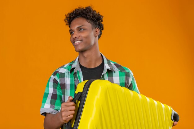 Jonge Afro-Amerikaanse reiziger man met koffer opzij glimlachend gelukkig en positief staande over oranje achtergrond kijken