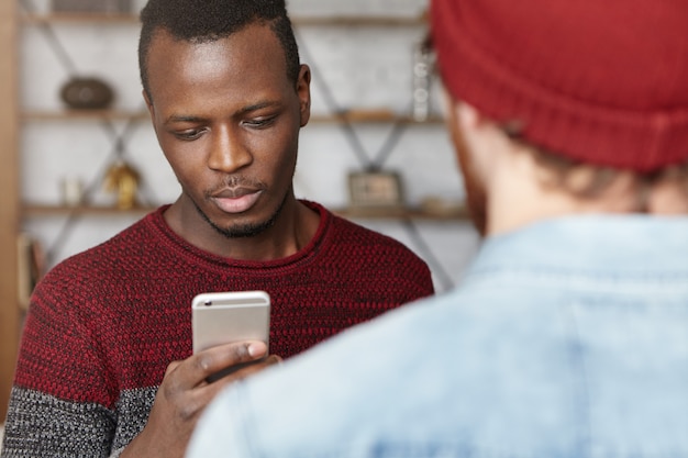 Jonge afro-amerikaanse student met mobiele telefoon, het typen van berichten tijdens een gesprek met zijn onherkenbare stijlvolle blanke vriend in de coffeeshop