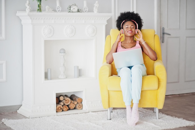 Jonge afro-amerikaanse vrouw die in een gele stoel zit terwijl ze op een laptop werkt en muziek luistert op oortelefoons
