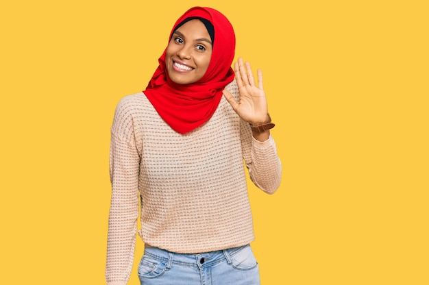 Gratis foto jonge afro-amerikaanse vrouw die traditionele islamitische hijab-sjaal draagt en afziet van hallo, gelukkig en glimlachend vriendelijk welkomstgebaar