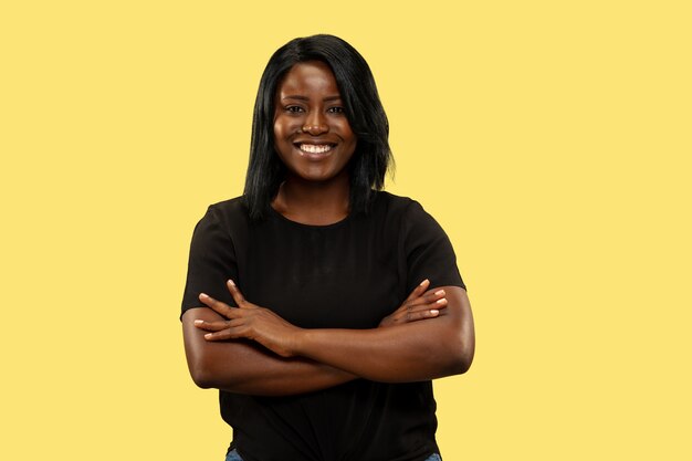 Jonge Afro-Amerikaanse vrouw geïsoleerd op gele studio achtergrond, gezichtsuitdrukking. Mooi vrouwelijk portret van halve lengte. Concept van menselijke emoties, gezichtsuitdrukking. Staande kruisende handen.