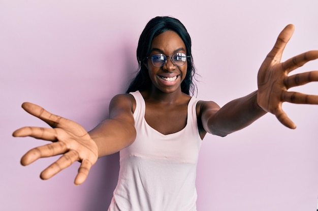 Gratis foto jonge afro-amerikaanse vrouw, gekleed in casual kleding en een bril, kijkend naar de camera glimlachend met open armen voor een knuffel, vrolijke uitdrukking die geluk omarmt