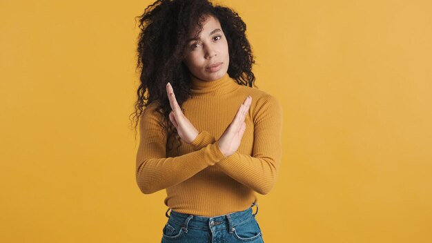 Jonge Afro-Amerikaanse vrouw met donker pluizig haar die er zelfverzekerd uitziet en de handen gekruist houdt en nee op de camera toont over een kleurrijke achtergrond