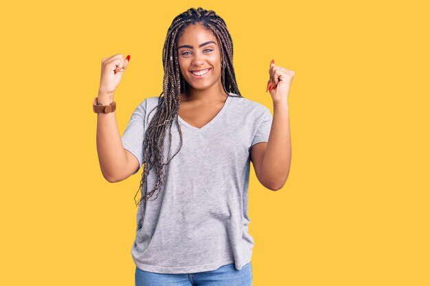Jonge afro-amerikaanse vrouw met vlechten die vrijetijdskleding dragen en trots schreeuwen om overwinning en succes te vieren, erg opgewonden met opgeheven armen