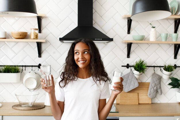 Jonge afro vrouw houdt twee glazen met water en melk en denkt welke keuze te maken