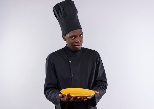 Jonge angstige afro-amerikaanse kok in uniform chef houdt plaat op wit met kopie ruimte