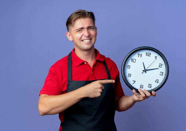 Jonge angstige blonde mannelijke kapper in uniforme ruimen en punten op klok geïsoleerd op violette ruimte met kopie ruimte