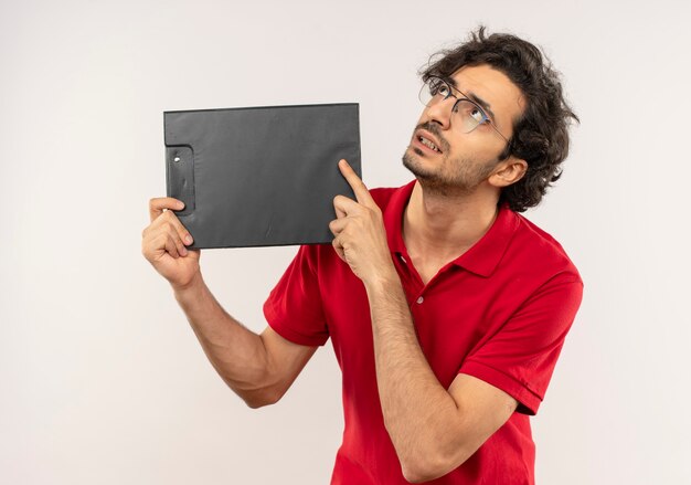 Jonge angstige man in rood shirt met optische bril houdt klembord en kijkt omhoog geïsoleerd op een witte muur