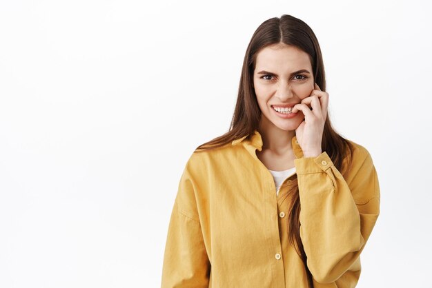 Jonge angstige vrouw die vingernagels bijt en bezorgd naar de camera kijkt, bang dat iets nerveus is tegen een witte achtergrond in een geel shirt