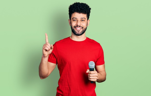 Jonge arabische man met baard zingt lied met behulp van microfoon glimlachend met een idee of vraag wijzende vinger met blij gezicht, nummer één