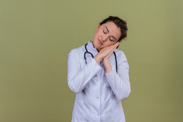 jonge arts medische jurk dragen stethoscoop toont slaap gebaar op groene muur