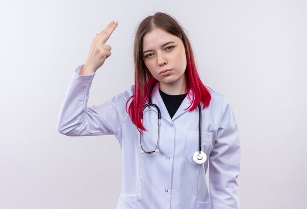 jonge artsenvrouw die stethoscoop medisch kleed draagt dat pistoolgebaar op geïsoleerde witte muur toont