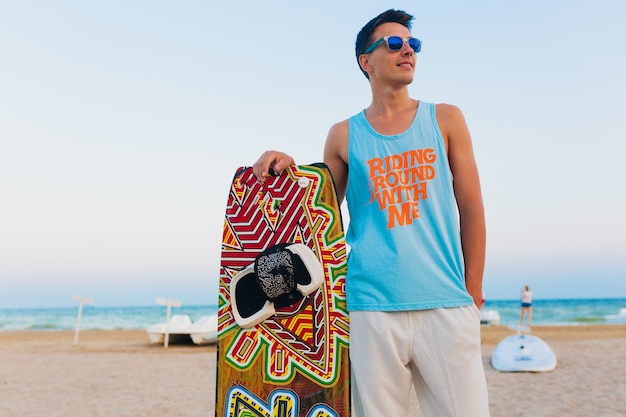 Gratis foto jonge atletische man met kitesurfplank die zich voordeed op het strand met een zonnebril op zomervakantie