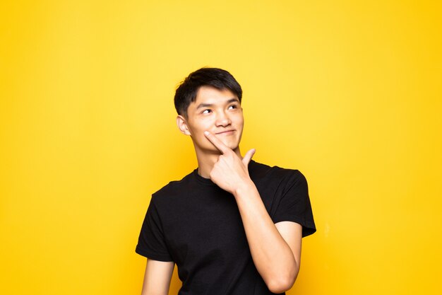 Jonge Aziatische Chinese mens die met hand op kin over vraag denkt, peinzende uitdrukking die zich over geïsoleerde gele muur bevindt. Glimlachend met een bedachtzaam gezicht. Twijfel concept.