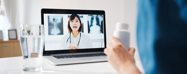 Jonge Aziatische dame die computerlaptop gebruikt, praat over een ziekte in een videoconferentiegesprek met een online consultatie van een senior arts in de woonkamer thuis.