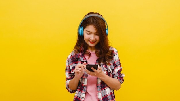 Jonge Aziatische dame die draadloze hoofdtelefoons dragen die aan muziek van smartphone met vrolijke uitdrukking in vrijetijdskleding luisteren en camera over gele muur bekijken. Gelaatsuitdrukking concept.