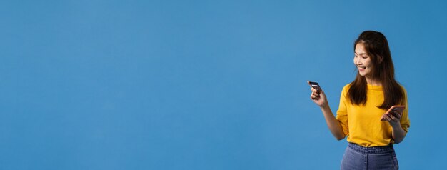Jonge Aziatische dame die telefoon en creditcard met positieve uitdrukking gebruikt, glimlach breed, gekleed in vrijetijdskleding en sta geïsoleerd op blauwe achtergrond. Panoramische bannerachtergrond met exemplaarruimte.