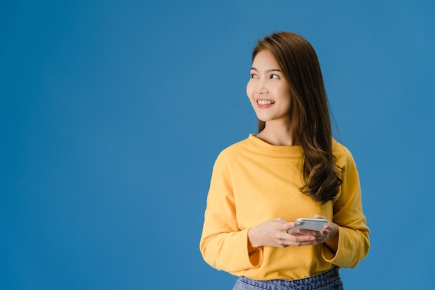 Jonge Aziatische dame die telefoon met positieve uitdrukking gebruikt, breed glimlacht, gekleed in vrijetijdskleding geluk voelt en status geïsoleerd op blauwe achtergrond. Gelukkige schattige blije vrouw verheugt zich over succes.