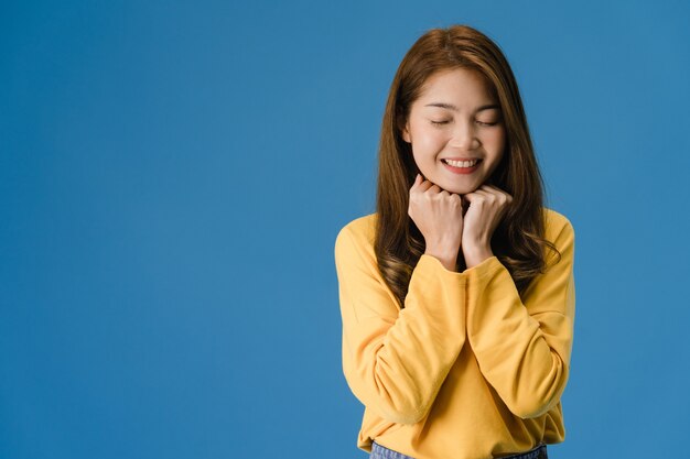 Jonge Aziatische dame met positieve uitdrukking, brede glimlach, gekleed in vrijetijdskleding en sluit je ogen op blauwe achtergrond. Gelukkige schattige blije vrouw verheugt zich over succes. Gezichtsuitdrukking concept.