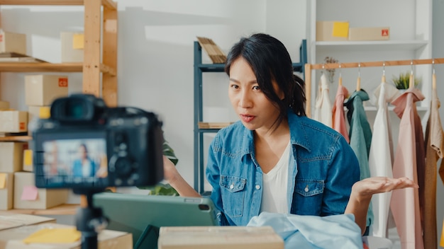 Jonge Aziatische dame modeontwerper met behulp van mobiele telefoon ontvangen inkooporder en kleding tonen in live streaming