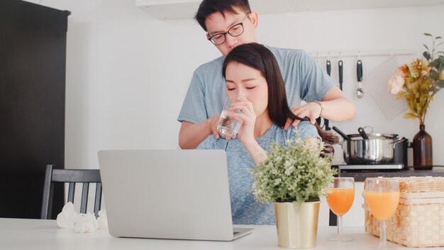 Jonge Aziatische ernstige onderneemster, spanning, vermoeid en ziek terwijl thuis het werken aan laptop. Man geeft haar glas water terwijl hij 's ochtends thuis hard aan het werk is in de moderne keuken.