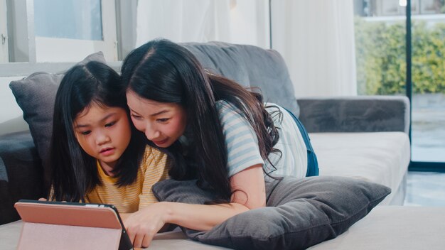 Jonge Aziatische familie en dochter gelukkige thuis gebruikende tablet. De Japanse moeder ontspant met meisje het letten op film liggend op bank in woonkamer bij huis. Grappige moeder en lief kind hebben plezier.