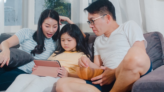 Jonge Aziatische familie en dochter gelukkige thuis gebruikende tablet. De Japanse moeder, vader ontspant met meisje het letten op film liggend op bank in woonkamer. De grappige ouder en het mooie kind hebben plezier.