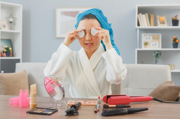 Jonge aziatische gelukkige vrouw met een handdoek op haar hoofd die thuis aan de kaptafel zit en ogen bedekt met wattenschijfjes die plezier heeft met het doen van ochtendmake-uproutine