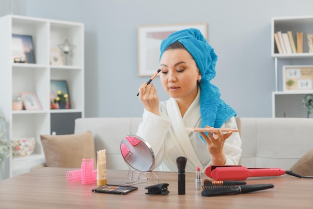 Jonge Aziatische gelukkige vrouw met handdoek op haar hoofd zittend aan de kaptafel thuis interieur oogschaduw toe te passen in spiegel kijken ochtend make-up routine schoonheid en gezichtscosmetica concept