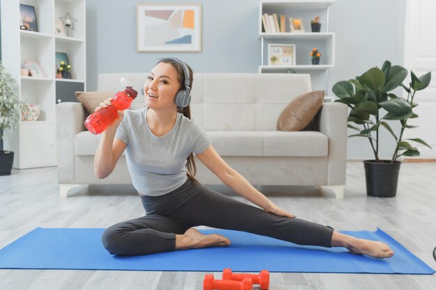 Jonge aziatische gezonde vrouw met koptelefoon die thuis oefeningen doet, haar benen strekkend om water te drinken in de woonkamer die op yogamat zit