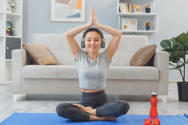 jonge Aziatische gezonde vrouw met koptelefoon doen oefening binnenshuis thuis mediteren in woonkamer zittend op yoga handpalmen bij elkaar houden boven haar hoofd glimlachen
