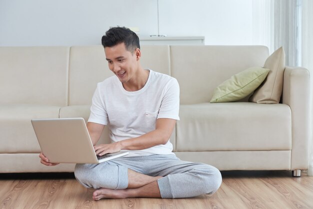 Jonge Aziatische kerel die zijn laptop op de vloer van de woonkamer naast de beige bank met behulp van