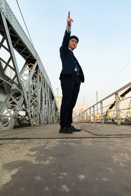 Jonge Aziatische man in een pak staande op een brug terwijl hij omhoog wijst