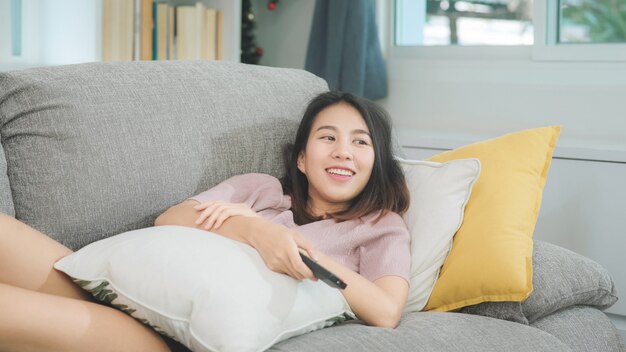 Jonge Aziatische tienervrouw die op TV thuis letten, wijfje die het gelukkige liggen voelen op bank in woonkamer. De levensstijlvrouw ontspant thuis in ochtendconcept.