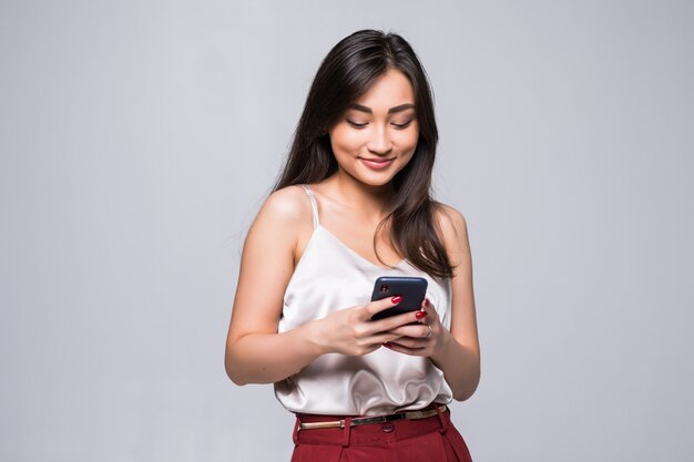 Jonge Aziatische vrouw die een slimme telefoon met behulp van die op witte muur wordt geïsoleerd.