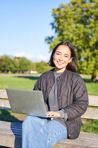 Jonge aziatische vrouw die op afstand werkt freelance meisje zit in het park met laptop die haar werk doet vanuit de buitenlucht