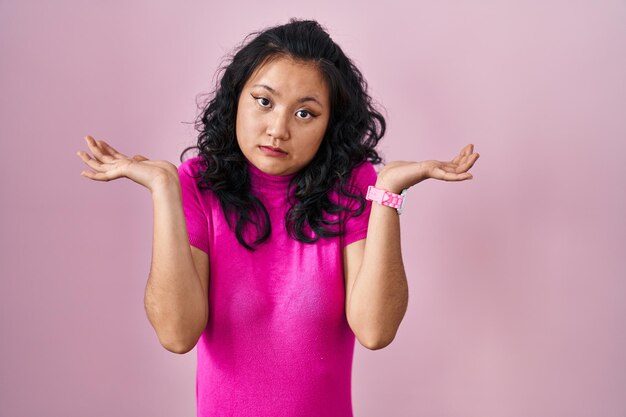 Jonge aziatische vrouw die over een roze achtergrond staat zonder idee en verwarde uitdrukking met armen en handen die twijfels opriepen