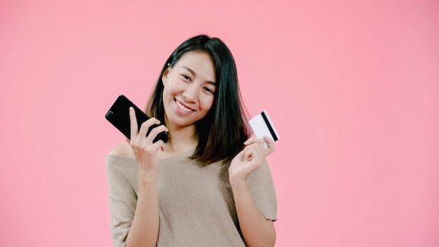 Jonge Aziatische vrouw die smartphone gebruiken die online winkelend door creditcard kopen die het gelukkige glimlachen in toevallige kleding over roze achtergrondstudioschot voelen.