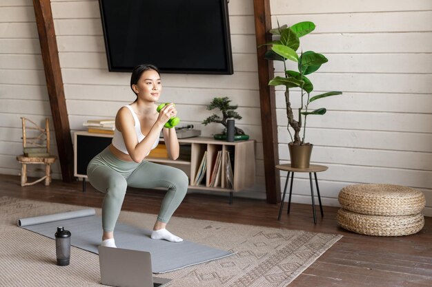 Jonge aziatische vrouw die thuis traint met dumbbels die squats op vloermat maken, volg online fitne...