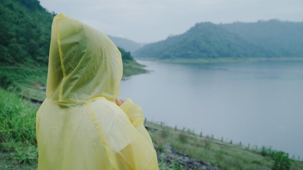 Jonge Aziatische vrouw die zich gelukkig voelt als ze regen speelt terwijl ze een regenjas draagt die in de buurt van het meer staat