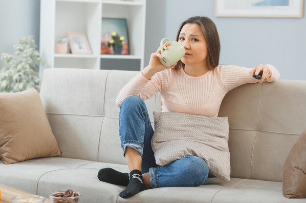 Jonge aziatische vrouw in vrijetijdskleding zittend op een bank in het interieur met afstandsbediening televisie kijken met sceptische uitdrukking die thee drinkt uit mok