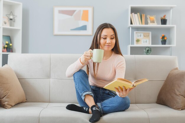 Jonge aziatische vrouw in vrijetijdskleding zittend op een bank thuis interieur met mok thee leesboek gelukkig en positief ontspannend weekend thuis