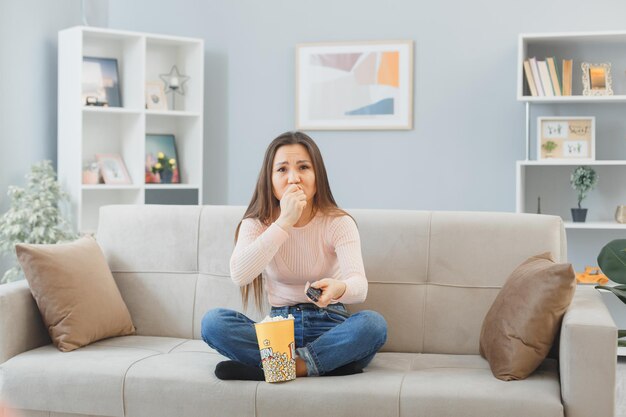 Jonge aziatische vrouw in vrijetijdskleding zittend op een bank thuis met een emmer popcorn die op afstand televisie kijkt, gestresst en nerveus