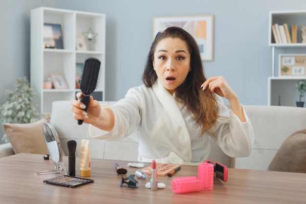 Jonge aziatische vrouw met een handdoek in een badjas die aan de kaptafel zit en haar haar borstelt en haar haarborstel laat zien die verward en overstuur is tijdens het doen van ochtendmake-uproutine